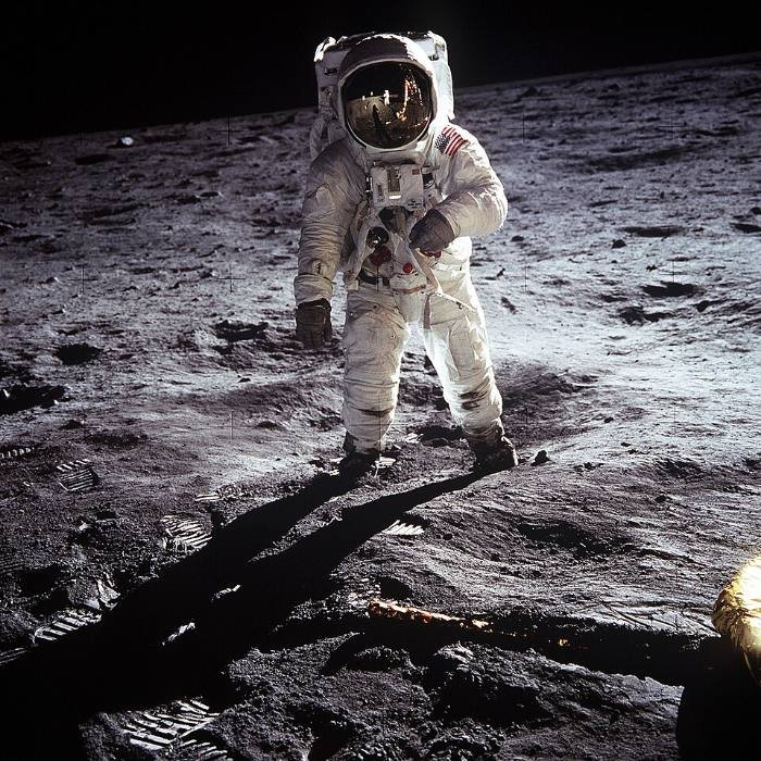 Ein Astronaut im Raumanzug steht auf der Mondoberfläche, im Visier spiegeln sich die Landefähre und der Fotograf